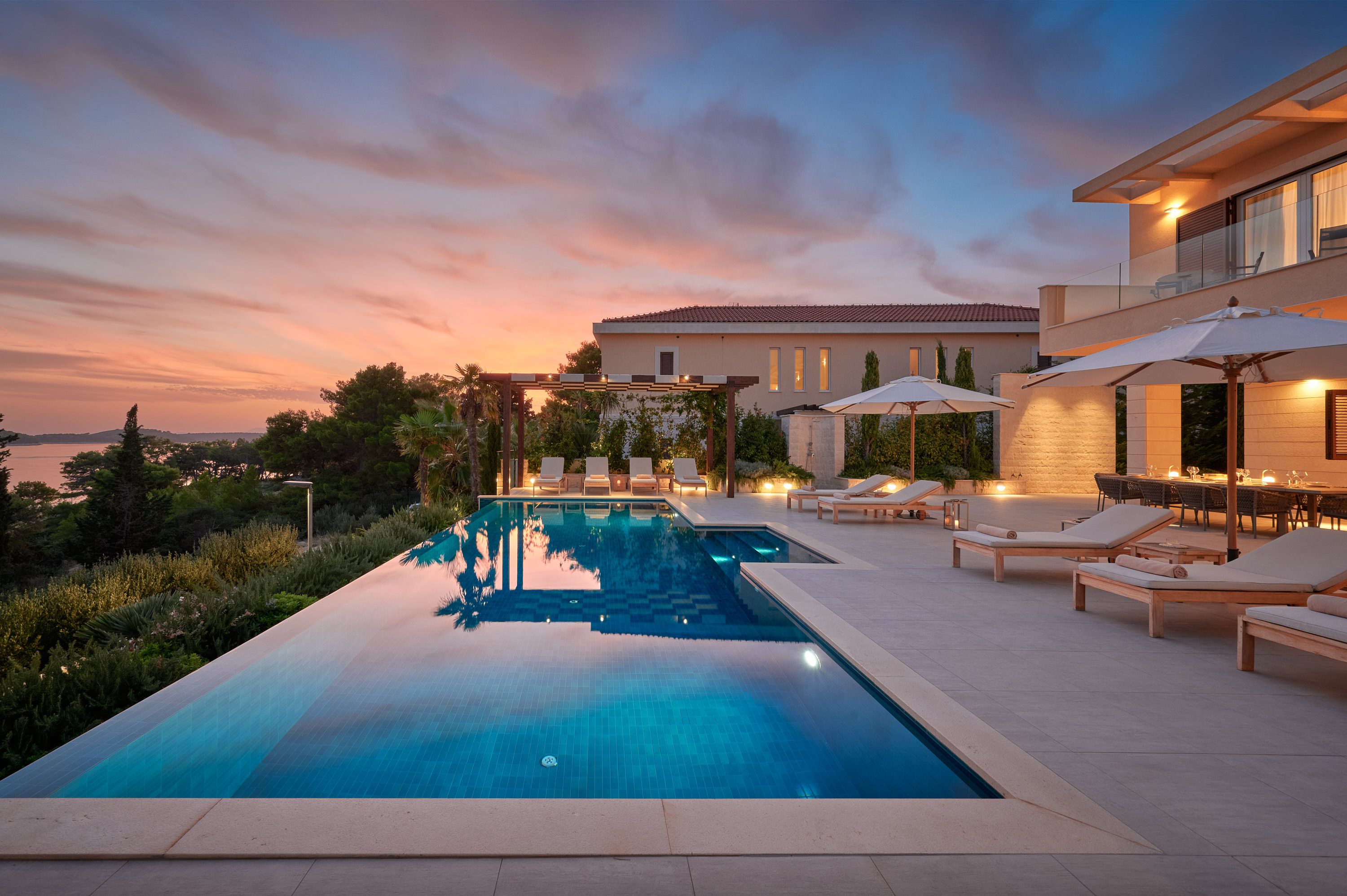 A luxurious villa Sapphire Hvar with an infinity pool at sunset, surrounded by lounge chairs and lush greenery.