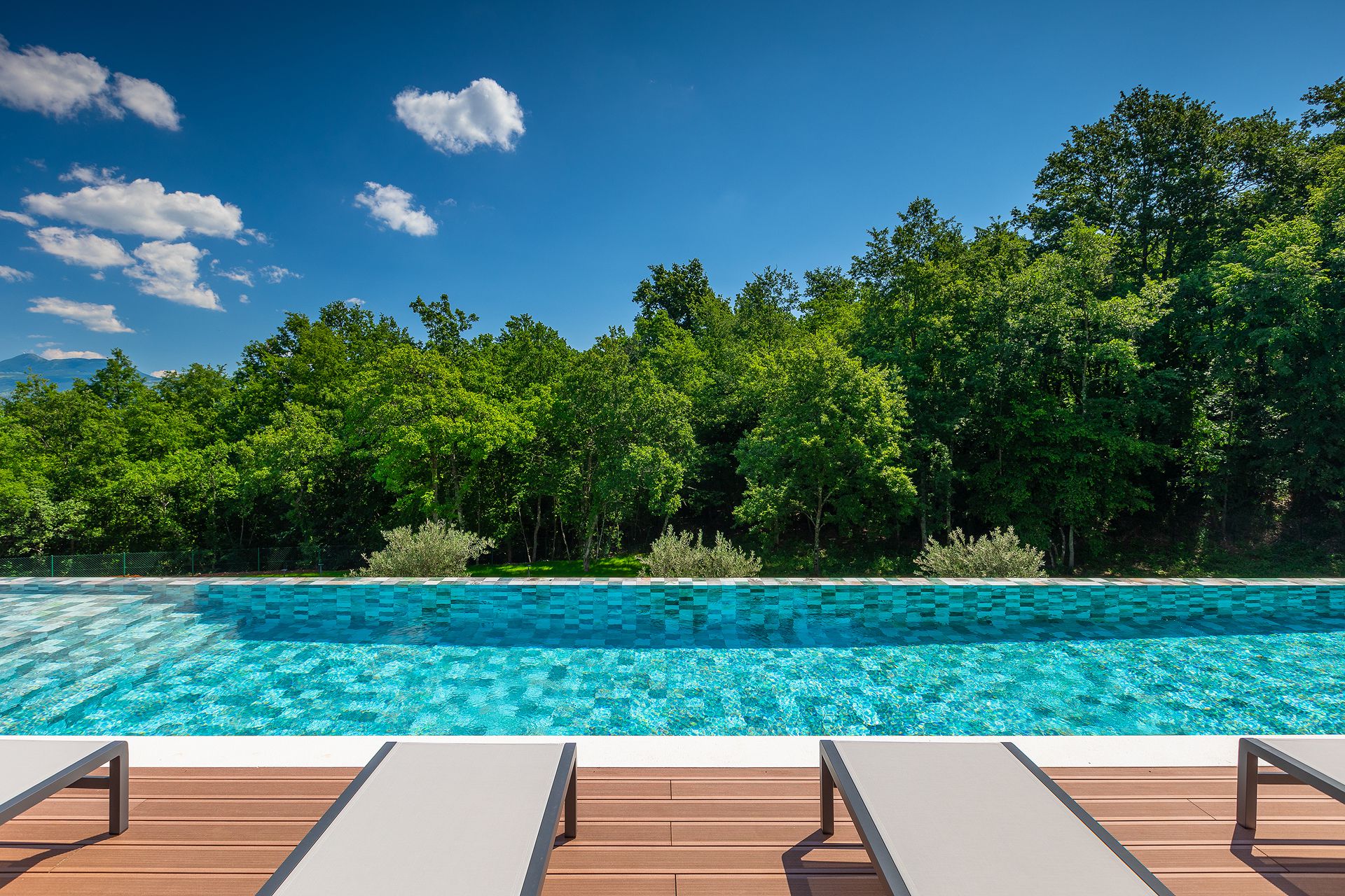 A swimming pool in Villa Chloe Istria with lounge chairs and a view of green trees under a blue sky.